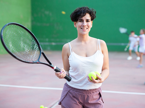 Portrait of positive latin woman frontenis player standing on outdoor fronton, holding racket and tennis ball, smiling and looking at camera.