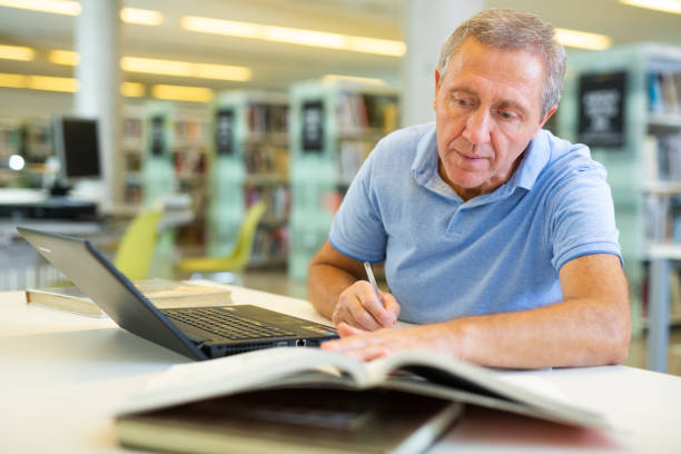 istock Mature man spending time in library, reading books and making laptop. Self-education concept 1481494293