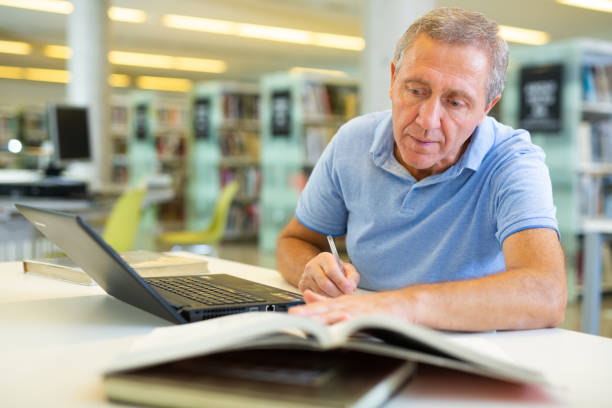 mature man spending time in library, reading books and making laptop. self-education concept - attentively imagens e fotografias de stock