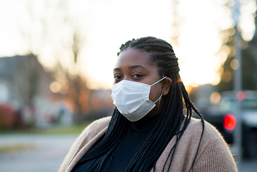 Beautiful young black woman is wearing a white protective face mask to prevent the spread of covid-19 virus or other illness to herself or others. She is standing outdoors on the sidewalk next to the street as she is waiting for a bus to arrive.