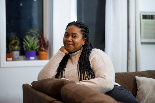 Young black woman with her hair in braids is smiling and sitting comfortably on a brown sofa in her living room and smiling at the camera in the evening.