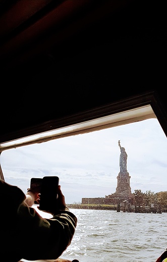 Close-up on a person's hand taking a photo of the Statue of Liberty on a boat tour. A second cell phone can also be seen in the back.