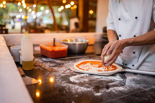 Italian male chef adding cheese on pizza dough in restaurant kitchen.