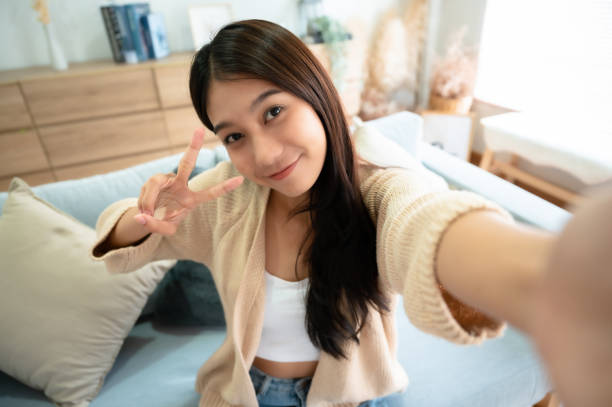 Pretty young asian female with big smile sitting at living room. She having fun taking light cheerful selfie on blurred background stock photo