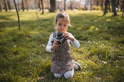 Portrait of an adorable little girl sitting on the grass in the park and taking pictures of nature with an instant camera