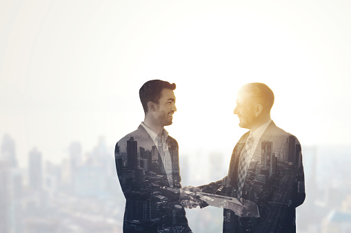 Double exposure of two businessmen handshaking together while standing near window with cityscape background