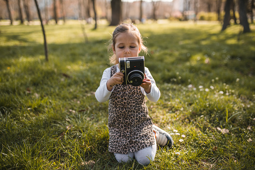 Portrait of an adorable little girl sitting on the grass in the park and taking pictures of nature with an instant camera