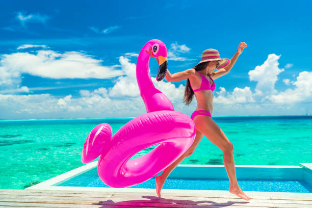 Vacation fun woman in bikini with funny inflatable pink flamingo pool float running of joy jumping by infinity swimming pool. Girl enjoying travel holidays at resort luxury overwater bungalow travel stock photo