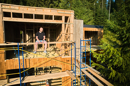 Designer is working on wooden frame two-story house construction site near the forest, sitting on scaffold. The concept behind it is modern and environmentally friendly construction.