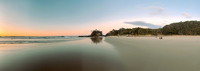 Horizontal landscape panoramic view of  sunset sky colours reflecting on wet sand at the water’s edge, Fisherman’s Lookout and coastal forest headland on the horizon at The Pass beach, Byron Bay, subtropical north coast of NSW.