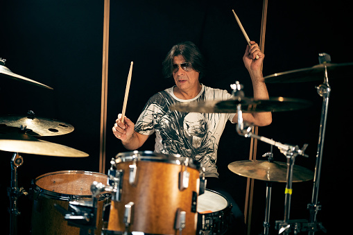 Mid-adult Caucasian male musician, with long dark hair and sunglasses playing drums, at the recording studio