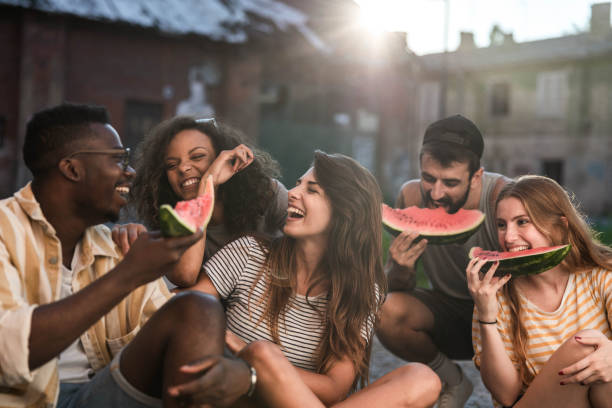 Young cheerful friends having fun while eating watermelon in nature. Group of cheerful friends having fun while eating fresh watermelon in the park. black man eating watermelon stock pictures, royalty-free photos & images