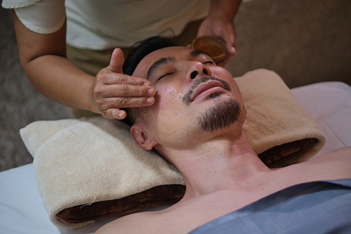 Hands of cosmetologist making manual relaxing rejuvenating facial massage for young asian man in beauty salon. Rejuvenating and beauty