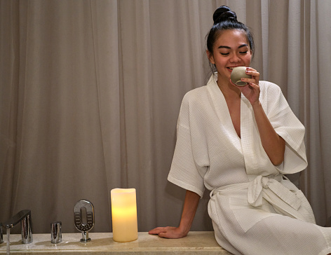 Young asian woman in white bathrobe, holding a cup of hot healthy herbal drink sitting beside bathtub surrounded by candle lights, relaxing and enjoying beauty procedures in spa salon