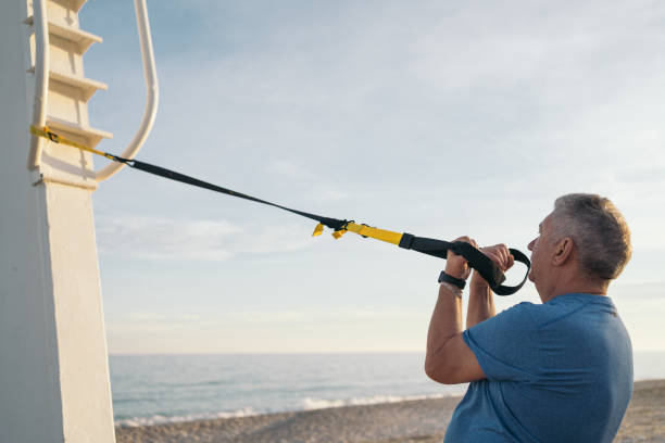 istock Side image of an older white-haired person with contracted arms performing biceps strengthening exercises from her suspension training on the beach at sunset. 1481604641