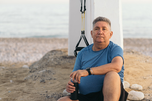 Overweight older white-haired man sitting on the sand of the beach clutching his water bottle looking exhausted after having done his suspension training.
