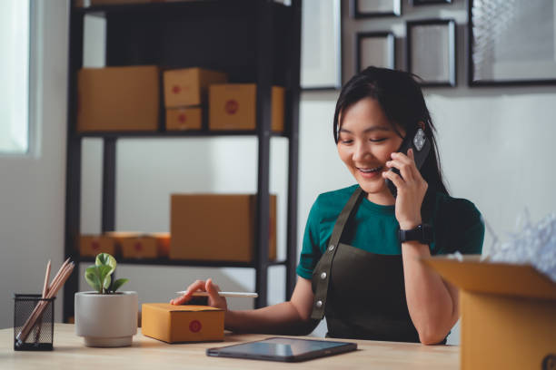istock Asian woman online seller using smartphone calling to customer confirming orders and make a note on tablet working at home office. 1481608356