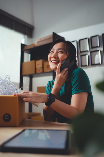 Asian woman online seller using smartphone calling to customer confirming orders and packing product for delivering. Small business owners working online at home.