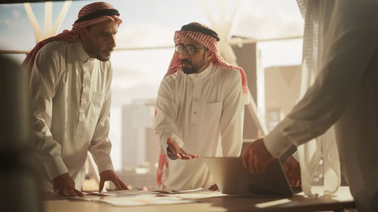 Successful Muslim Businessmen in White Traditional Outfits Having an Office Meeting, Negotiating and Talking About Financial Opportunities. Using Laptop. Saudi, Emirati, Arab Businessman Concept.