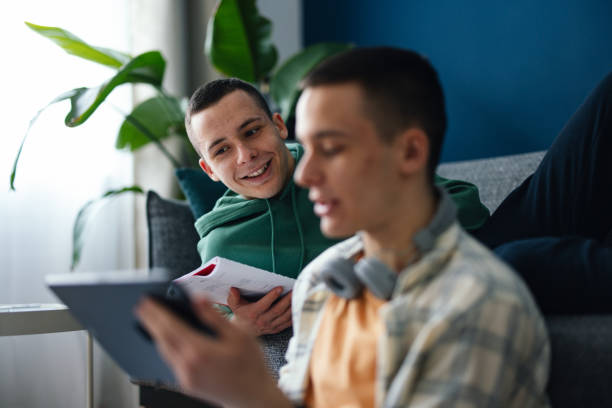 An Anonymous Teenager Using A Digital Tablet With His Twin Brother At Home Two teenage boys reading an online lesson on a tablet while sitting in the living room together. blue brothers stock pictures, royalty-free photos & images