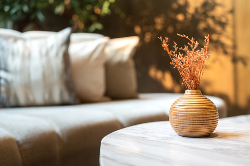 A cozy styled wooden flower pot with dry plant is placed on marble table with background of luxury sofa seating at luxury lounge. Interior decoration object photo, close-up and selective focus.