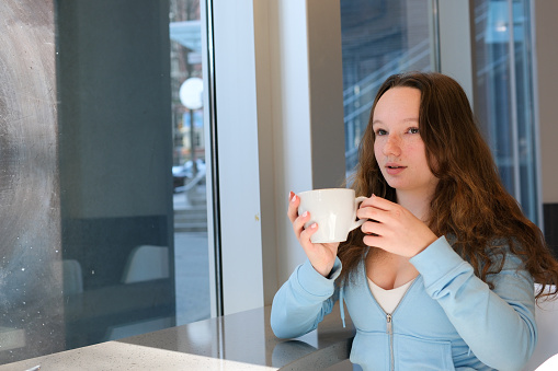 Close up portrait of a beautiful young woman drinking a cup of coffee by a window at home