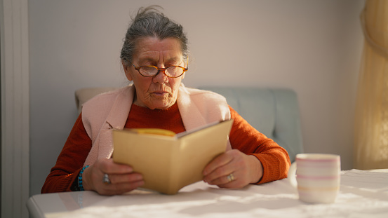 A senior woman is sitting at a table and enjoying reeding a book.