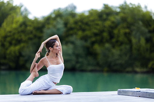 Young woman in white doing stretching exercises on a pier at lake. Copy space.