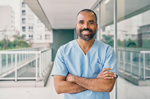 Portrait of confident mid adult male nurse standing in hospital. Smiling african healthcare worker wearing scrubs with arms crossed and looking at camera.
