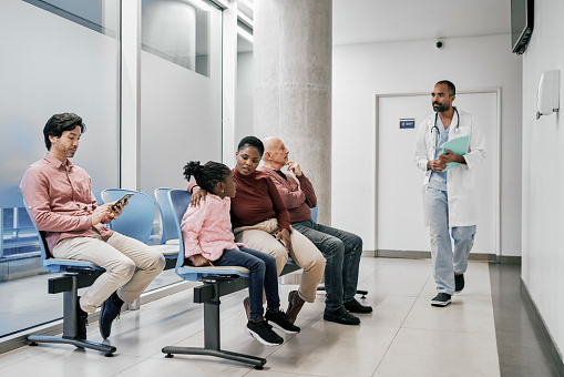 Group of people sitting in a waiting room of a hospital with a doctor arriving. Multiracial group of people wait for their appointment in a medical clinic.