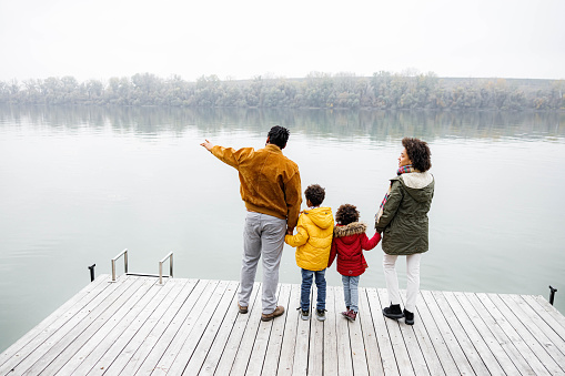 Back view of African American family holding hands on a jetty and looking at river view in winter day. Man is aiming at distance. Copy space.