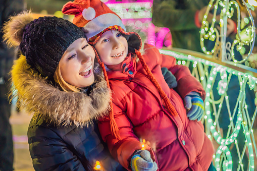 Little boy and his mother with sparklers near giant fir tree and Christmas illumination on Christmas market. Xmas holidays on fair.