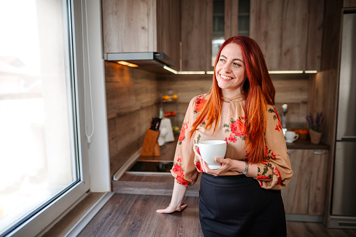 Smiling redhead woman looking through the kitchen window while drinking her morning coffee