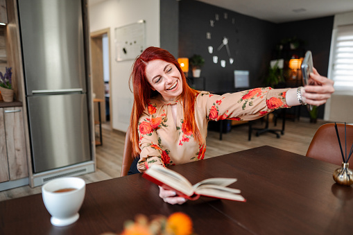 Smiling redhead woman taking selfies while reading a book in her apartment