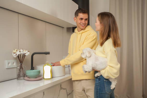 Portrait of happy couple in the kitchen, man washing the dishes, woman holding little white dog, dishwashing liquid with blank label on the table Portrait of happy couple in the kitchen, man washing the dishes, woman holding little white dog, dishwashing liquid with blank label on the table dog dishwasher stock pictures, royalty-free photos & images