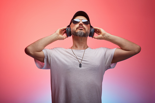 Smiling young man dancing to his favourite music in a studio. Happy young man enjoying streaming music with wireless earphones. Carefree young man standing alone against a pink background.