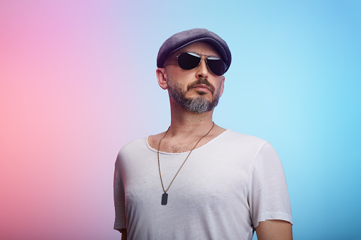 Studio portrait of a middle-aged confident man wearing a t-shirt, cap and sunglasses posing against a colorful background