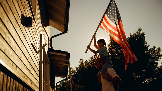 Cinematic Shot of a Father Holding His Small Daughter of His Shoulders, Helping Her to Raise the United States of America Flag to Celebrate a National Fourth of July Holiday at Their House.