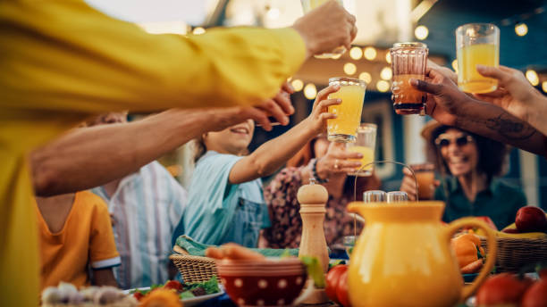 famiglia e amici diversi multiculturali che si riuniscono in una cena in giardino. vecchi e giovani alzano e tintinnano bicchieri con succo d'arancia fresco e celebrano l'occasione. - front or back yard house family barbecue foto e immagini stock