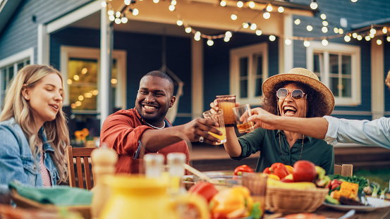 Family and Multiethnic Diverse Friends Gathering Together at a Garden Table Dinner. Old and Young People Toasting and Clinking Glasses with Fresh Orange Juice and Celebrating an Event.