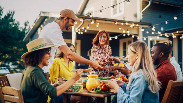 istock Handsome Man Cooking Grilled Corn and Other Vegetables. Bringing Food to a Big Outdoors Table with Family Members and Friends. Diverse Group of Adults and Children Having a Vegetarian Dinner. 1481647498