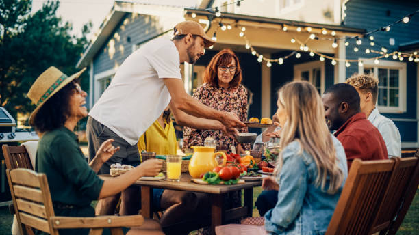 istock Handsome Man Cooking Grilled Corn and Other Vegetables. Bringing Food to a Big Outdoors Table with Family Members and Friends. Diverse Group of Adults and Children Having a Vegetarian Dinner. 1481647525