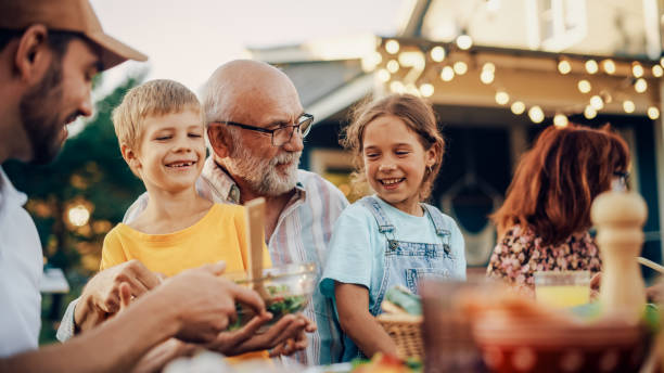 Happy Senior Grandfather Talking and Having Fun with His Grandchildren, Holding Them on Lap at a Outdoors Dinner with Food and Drinks. Adults at a Garden Party Together with Kids. Happy Senior Grandfather Talking and Having Fun with His Grandchildren, Holding Them on Lap at a Outdoors Dinner with Food and Drinks. Adults at a Garden Party Together with Kids. grandparent stock pictures, royalty-free photos & images