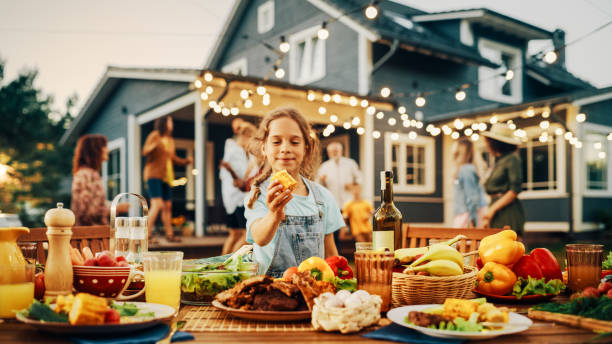 istock Outdoors Dinner Table with Delicious Barbecue Meat and Fresh Vegetables and Salads. Little Girl Eating a Grilled Corn. Happy People Dancing and Having Fun in the Background. 1481647675