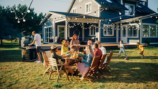 Parents, Children and Friends Gathered at a Barbecue Dinner Table Outside a Beautiful Home. Old and Young People Have Fun, Eat and Drink. Garden Party Celebration in a Backyard.