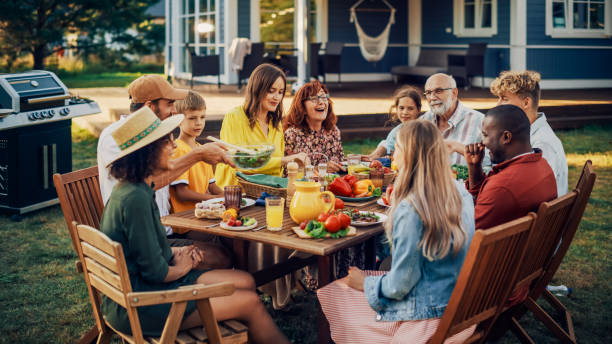 pais, filhos, parentes e amigos tendo um jantar vegetariano ao ar livre em seu quintal. idosos e jovens conversam, conversam, se divertem, comem e bebem. celebração da festa no jardim em um quintal. - encontro social - fotografias e filmes do acervo