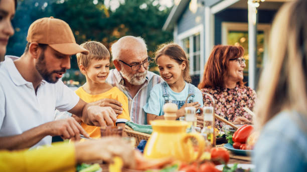 Portrait of a Happy Senior Grandfather Holding His Bright Talented Little Grandchildren on Lap at a Outdoors Dinner Party with Food and Drinks. Family Having a Picnic Together with Children. Portrait of a Happy Senior Grandfather Holding His Bright Talented Little Grandchildren on Lap at a Outdoors Dinner Party with Food and Drinks. Family Having a Picnic Together with Children. three generation family stock pictures, royalty-free photos & images