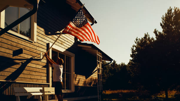 Proud American Homeowner Flying the United States of America National Flag on His Residential Area Home. Adult Man Fixing the Flag on His House Wall. Perfect Summer Day to Celebrate Patriotism. orgulloso propietario estadounidense que ondea la bandera nacional de los estados unidos de américa en su casa de área residencial. hombre adulto arreglando la bandera en la pared de su casa. día de verano perfecto para celebrar el patriotismo.