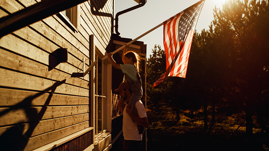 Proud Patriotic Dad Holding His Young Daughter of His Shoulders, Helping Her to Put the United States of America Flag on the Wall of Their House to Celebrate a National Holiday. Sunny Day in USA.