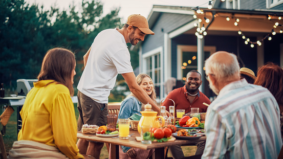 Young Adult Man Bringing Prepared Barbecue Meat Burgers to the Table with Young and Senior Relatives and Friends. Multicultural Group of Adults and Children Having an Outdoors Dinner Party.